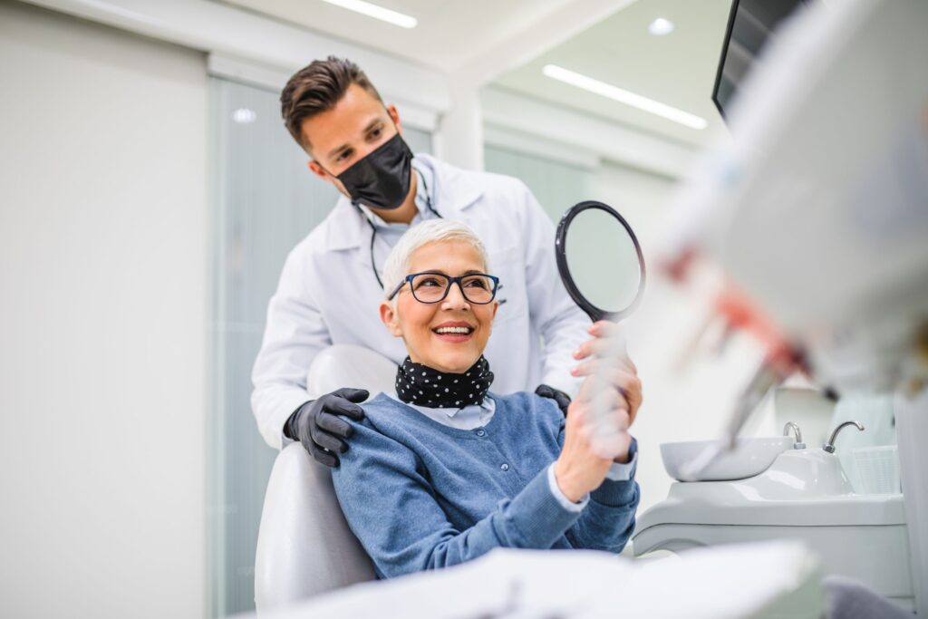 Woman with white hair smiling at reflection with dentist touching her shoulders
